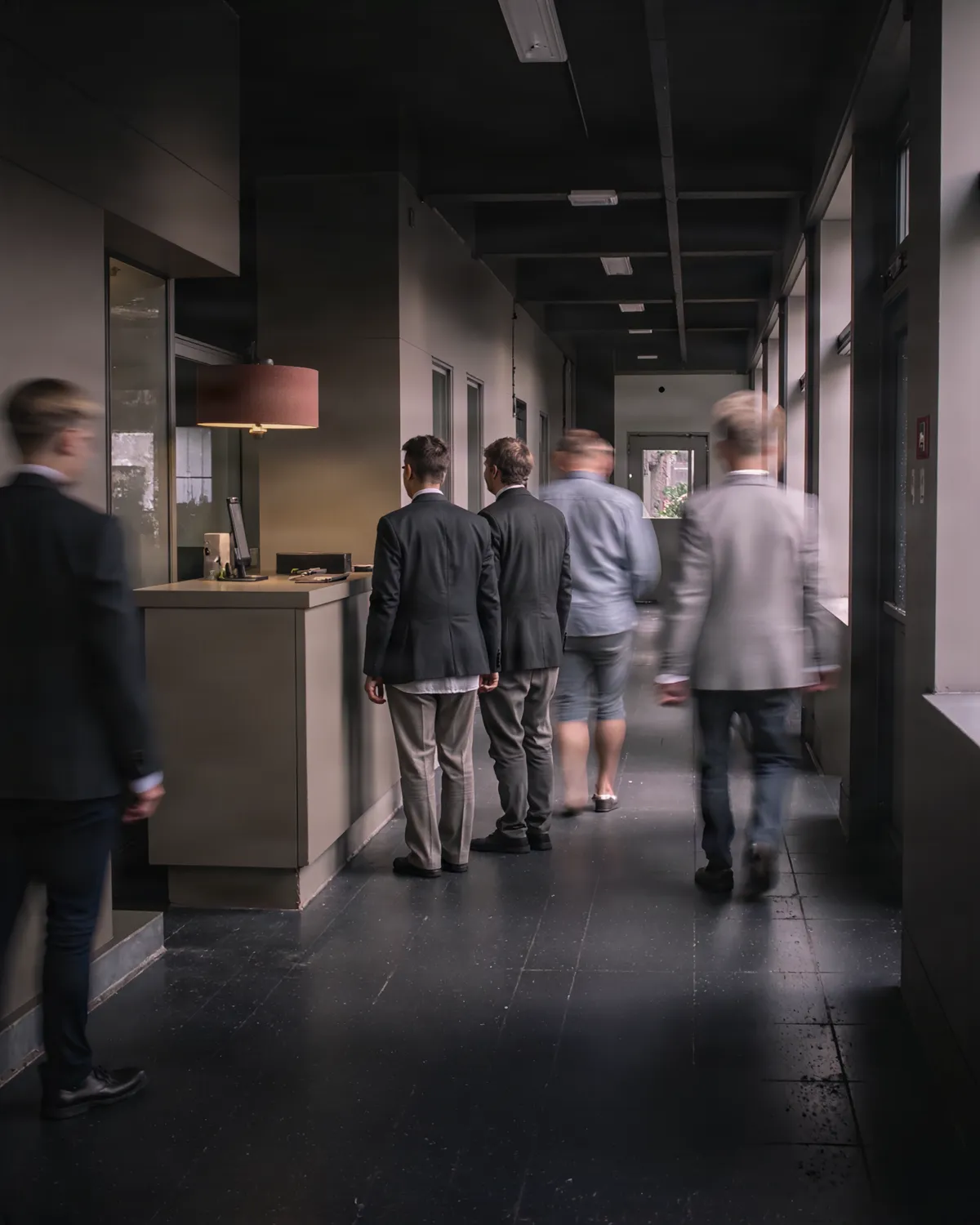 A corridor with a service counter. Passers-by blur in motion, only the three people waiting at the counter stand sharp.